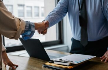 Two professionals shaking hands over a desk with documents and a laptop, symbolizing successful business agreement